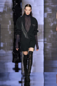 Model walking a runway in a black blazer with fringe detail over a dark turtleneck and thigh-high boots.