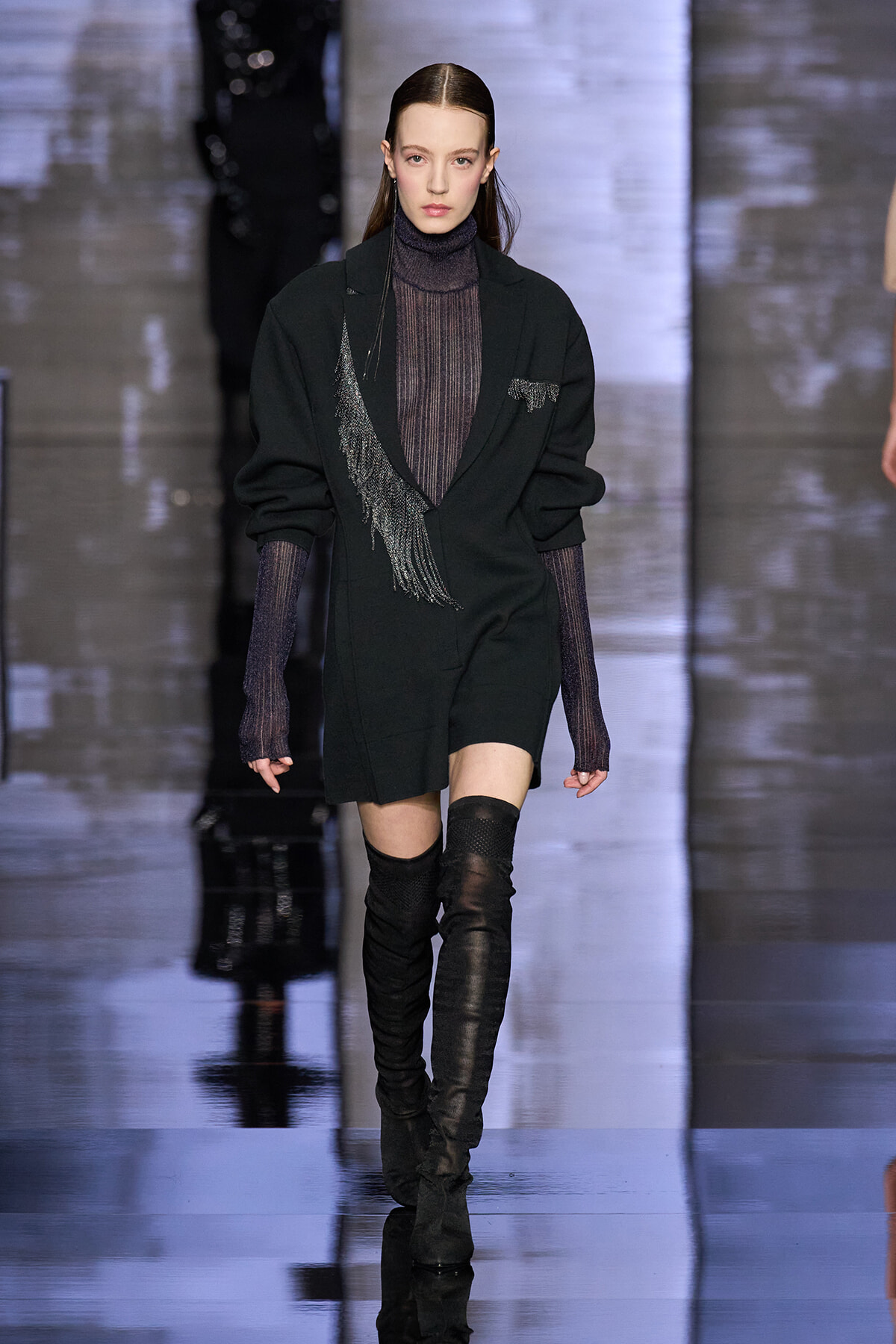 Model walking a runway in a black blazer with fringe detail over a dark turtleneck and thigh-high boots.
