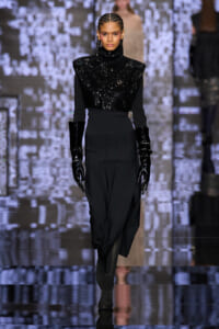 Model walking a runway in a black sequined turtleneck top, long black skirt, and shiny black gloves at a fashion show.
