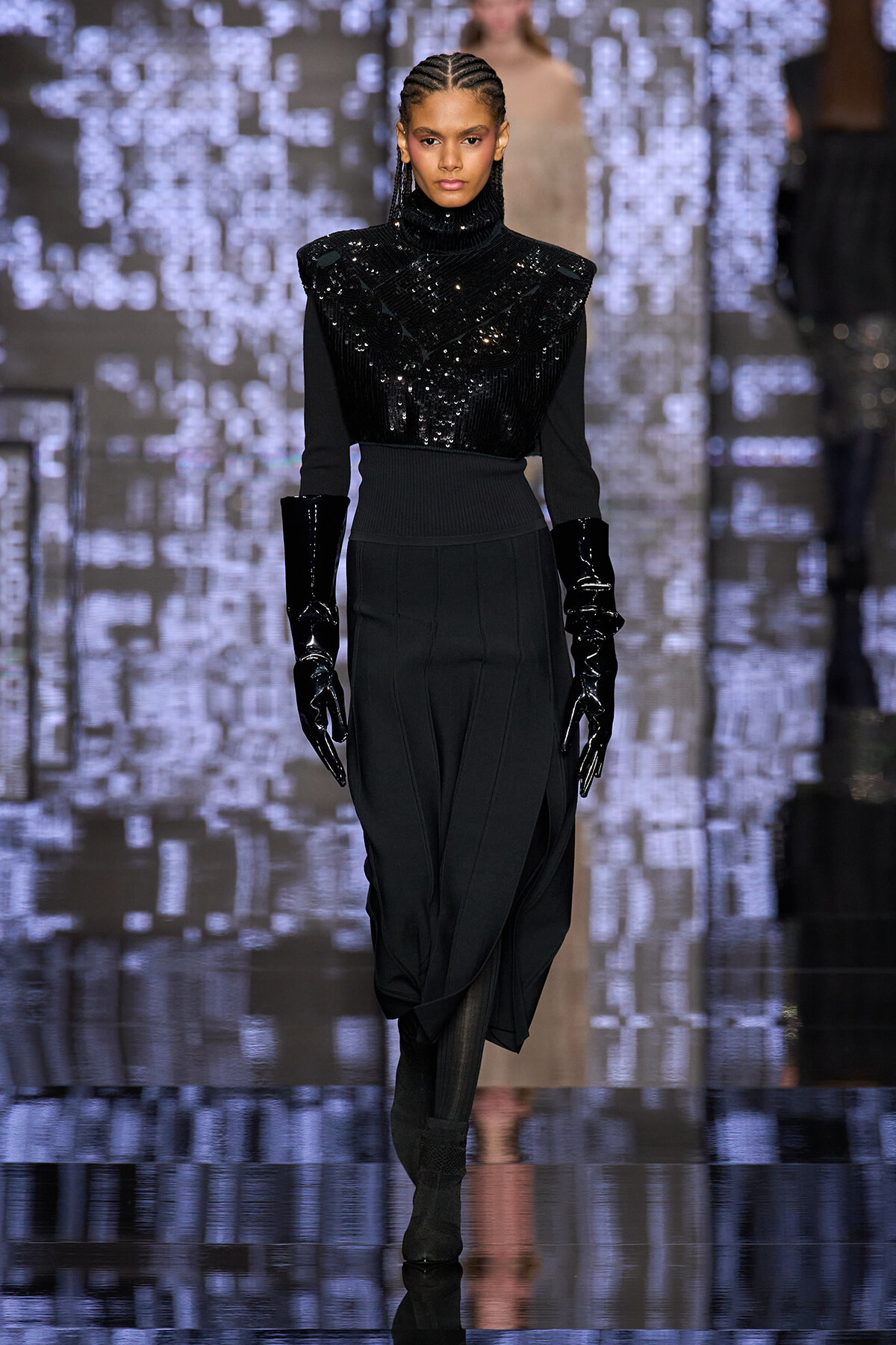 Model walking a runway in a black sequined turtleneck top, long black skirt, and shiny black gloves at a fashion show.