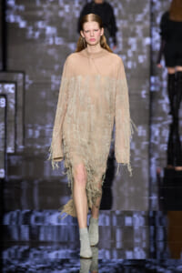 Model walking the runway in a beige, fringed, knee-length dress with long fringe accents on sleeves and skirt.