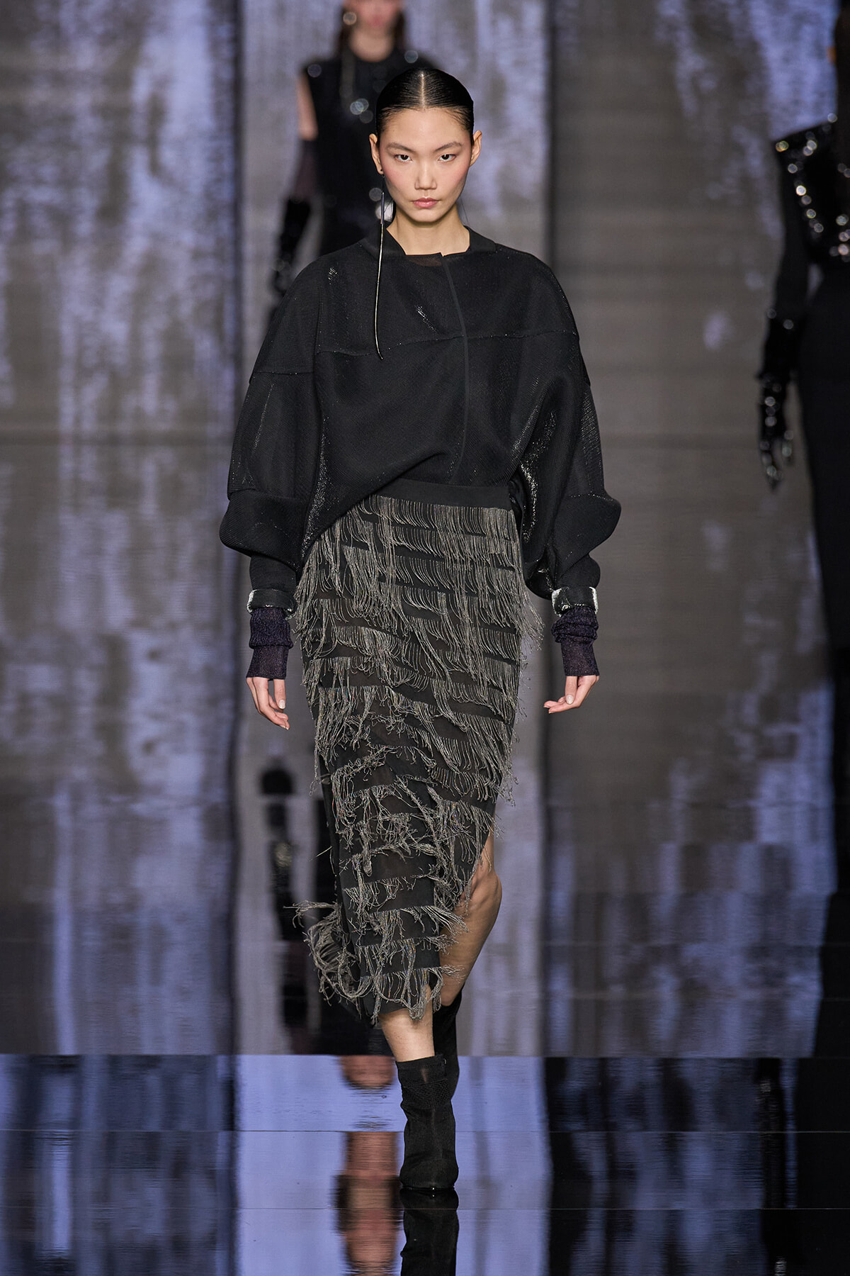 Model walks the runway in an oversized black sweater and a fringed gray-black skirt, slicked-back hair, mid-stride on a glossy catwalk.