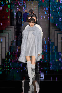 Female model walks a runway wearing a metallic silver coat over a light gray dress and over-the-knee glitter boots; colorful digital backdrop behind.