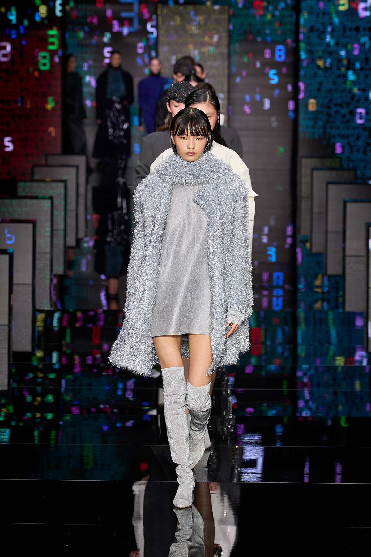 Female model walks a runway wearing a metallic silver coat over a light gray dress and over-the-knee glitter boots; colorful digital backdrop behind.