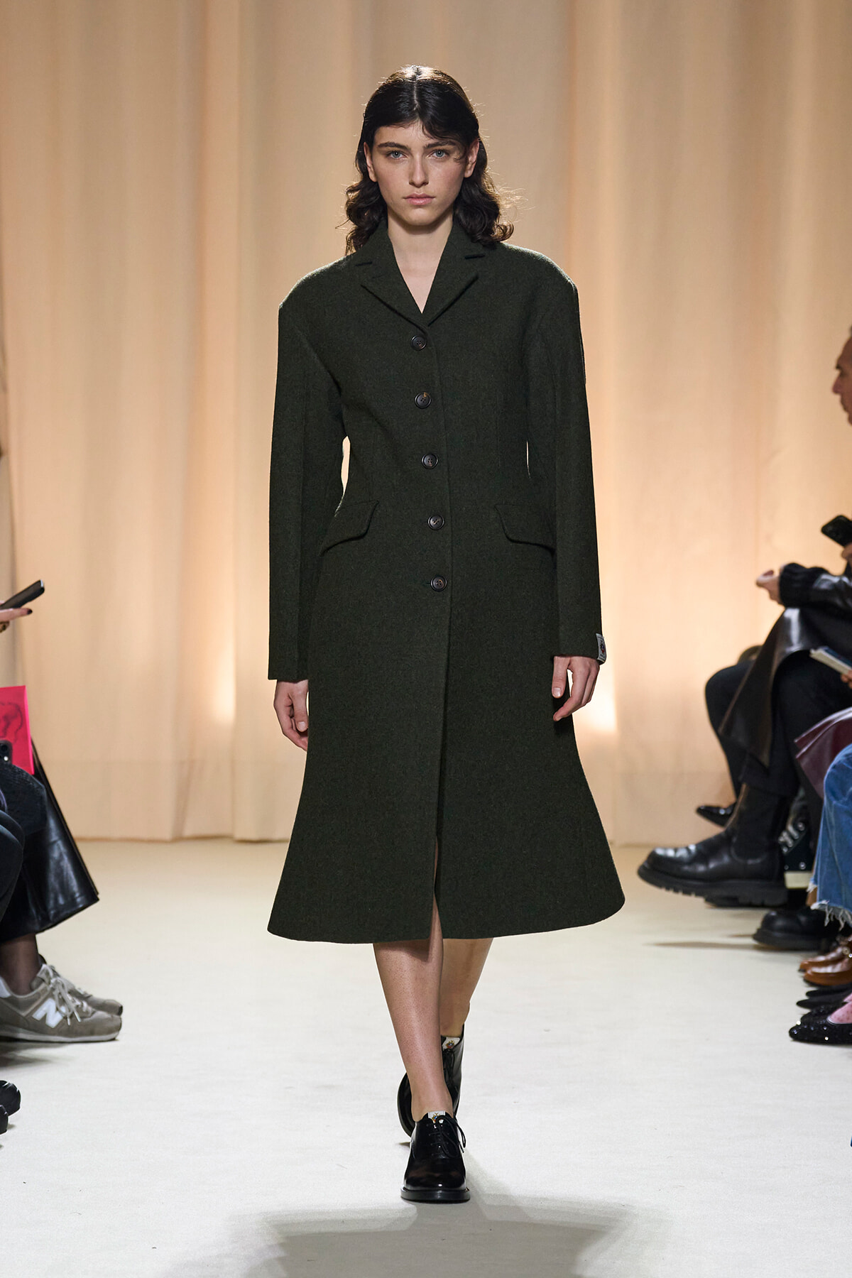 Model walks the runway wearing a dark green, knee-length, single-breasted wool coat with large buttons and pockets, paired with black shoes.