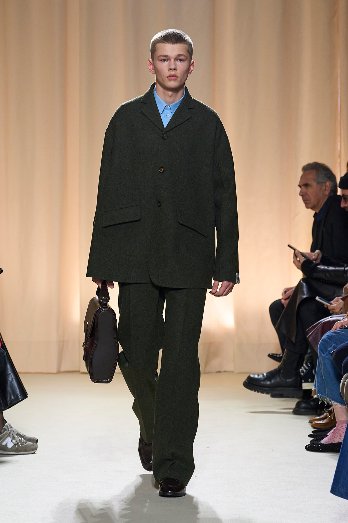 Male model walks a fashion runway in a dark green oversized suit, blue shirt, and polished shoes, carrying a brown briefcase.