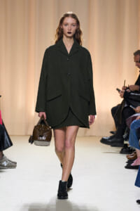 Model walking the runway in a dark green oversized coat, carrying a brown handbag and wearing black boots against a beige backdrop.