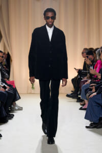 Male model walks a runway in a black velvet suit with a white shirt and sunglasses, holding a small sprig of greenery down the catwalk.