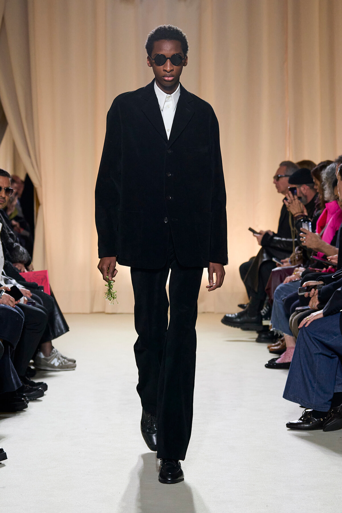 Male model walks a runway in a black velvet suit with a white shirt and sunglasses, holding a small sprig of greenery down the catwalk.