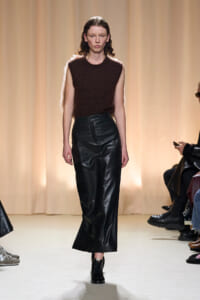 Model walks the runway in a sleeveless brown knit top and black leather wide-leg pants, beige curtain backdrop.