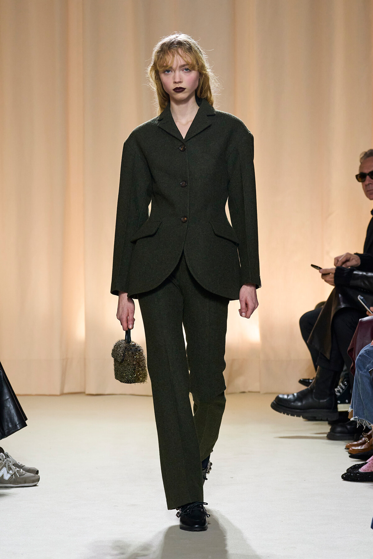 Female model walks the runway in a dark green tailored suit, holding a small textured handbag with a rounded shape.