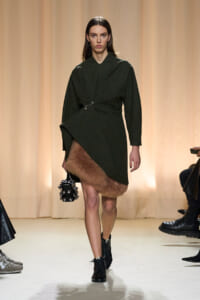 Model on a runway wearing a dark green wrap coat with a fur-trimmed hem, black lace-up boots, and a spiked handbag.