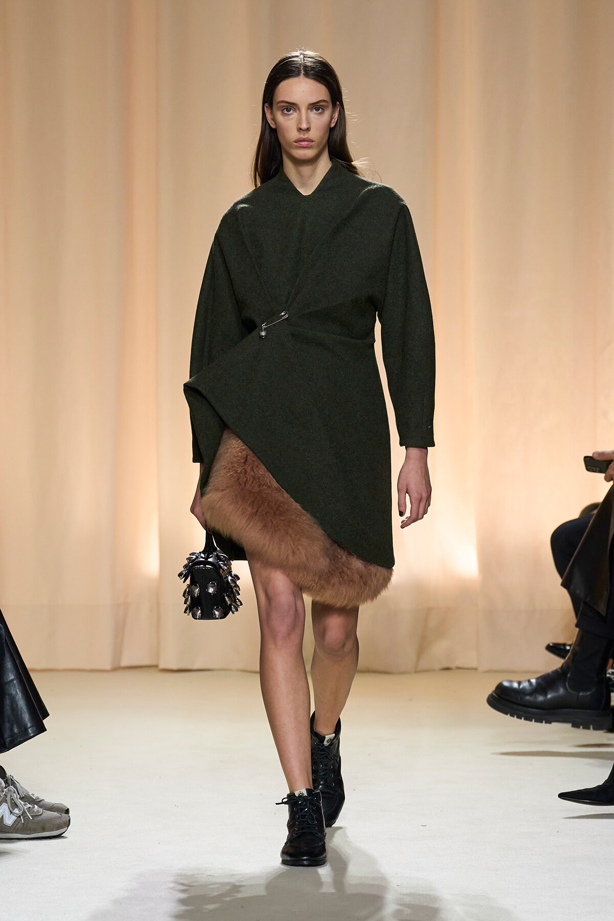 Model on a runway wearing a dark green wrap coat with a fur-trimmed hem, black lace-up boots, and a spiked handbag.
