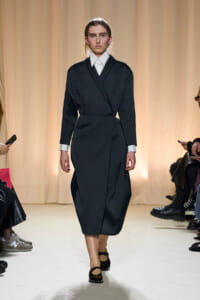 Model walking a fashion runway in a black wrap dress worn over a white collared shirt, beige backdrop behind.