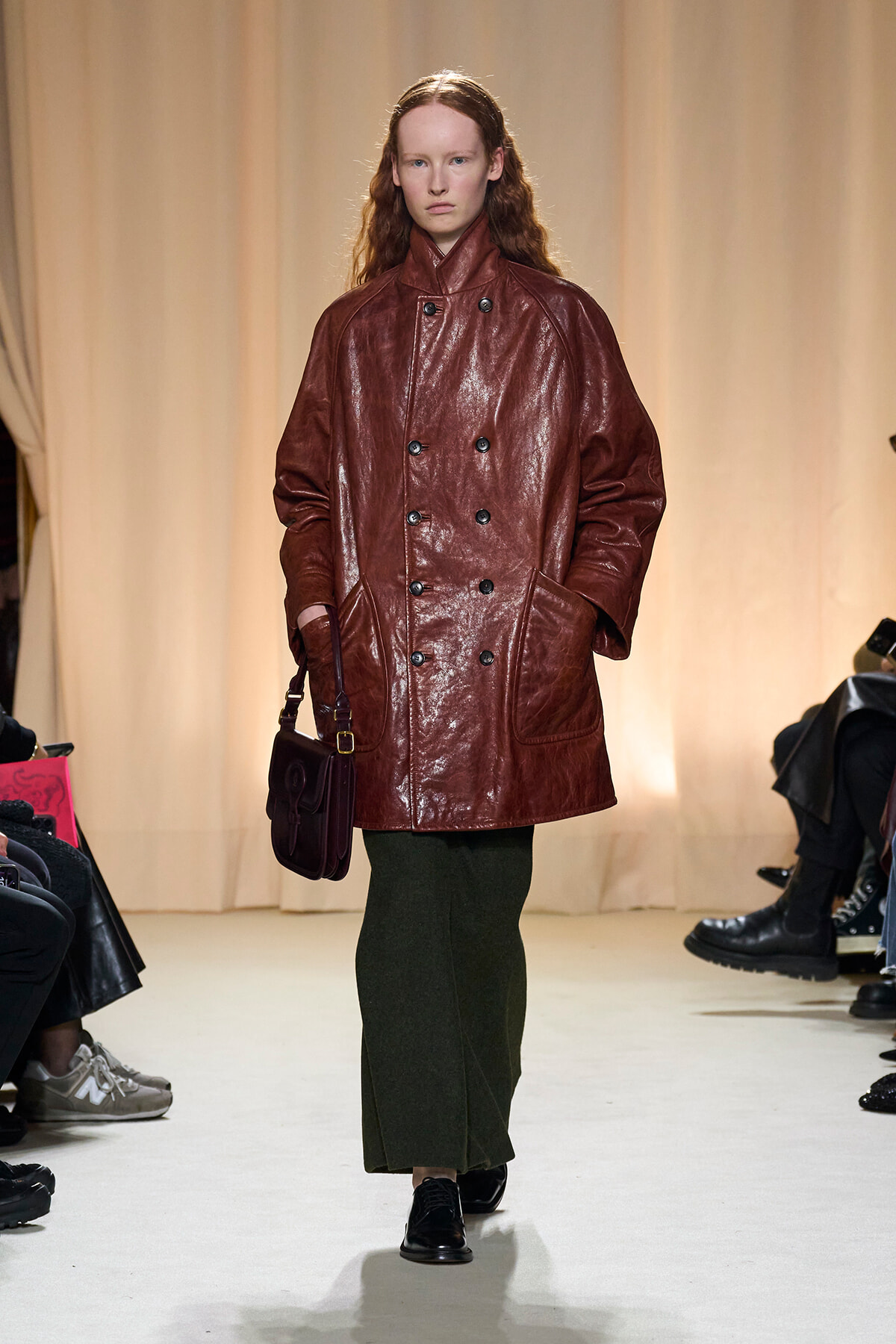 Model on a runway wearing a burgundy double-breasted leather coat, dark green skirt, and a small burgundy handbag.