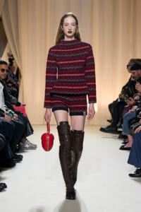 Model walks a runway in a red and black knit matching set, with tall brown lace-up boots and a red spherical handbag.