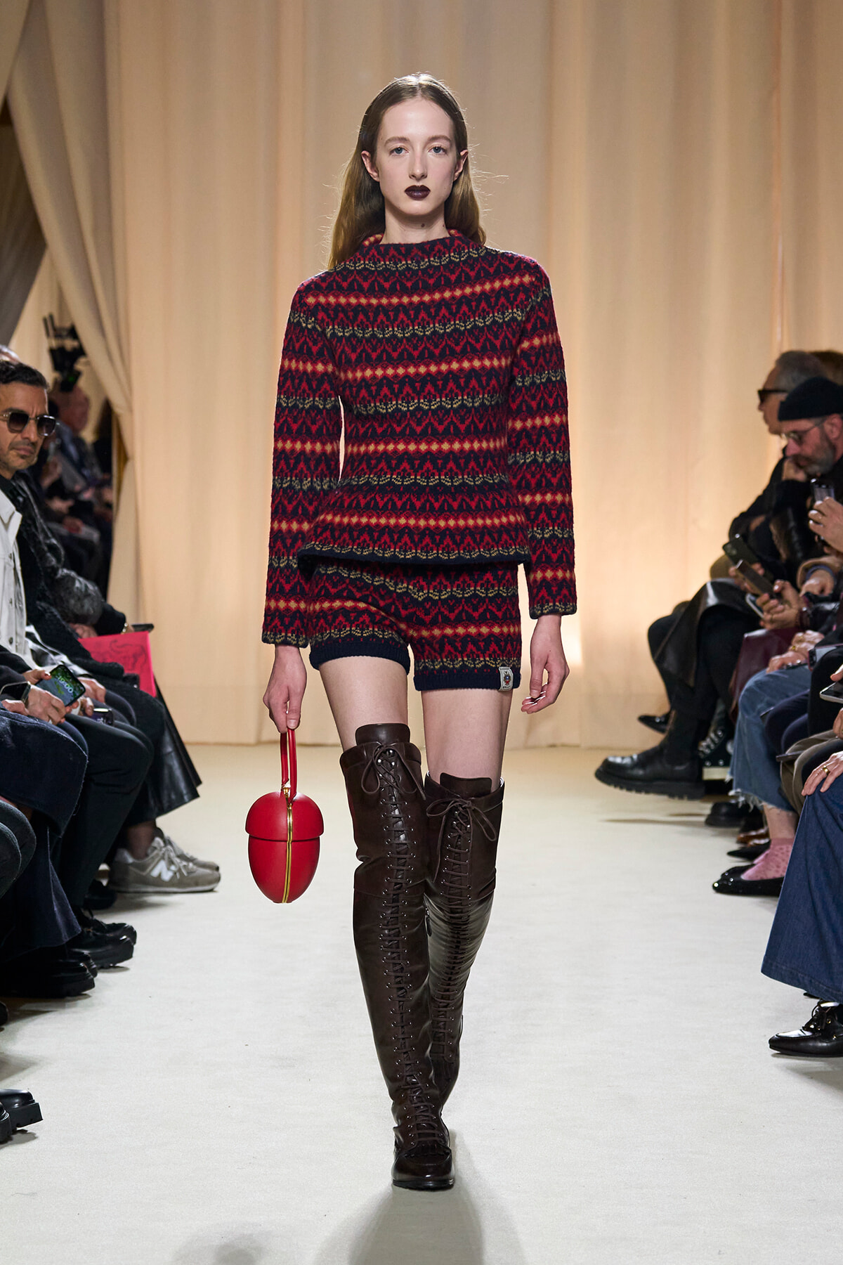 Model walks a runway in a red and black knit matching set, with tall brown lace-up boots and a red spherical handbag.