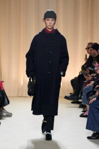 Model walking a fashion runway in a dark navy duffle coat with wooden toggles, black beanie, and a small black shoulder bag; audience seated along the runway.