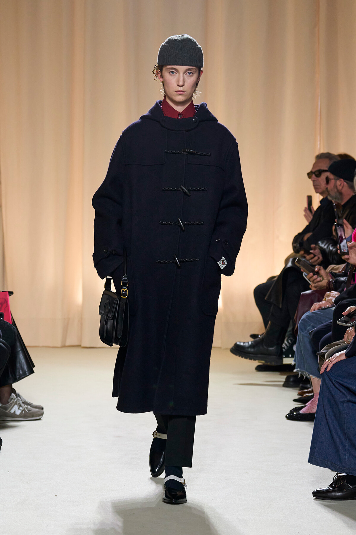 Model walking a fashion runway in a dark navy duffle coat with wooden toggles, black beanie, and a small black shoulder bag; audience seated along the runway.