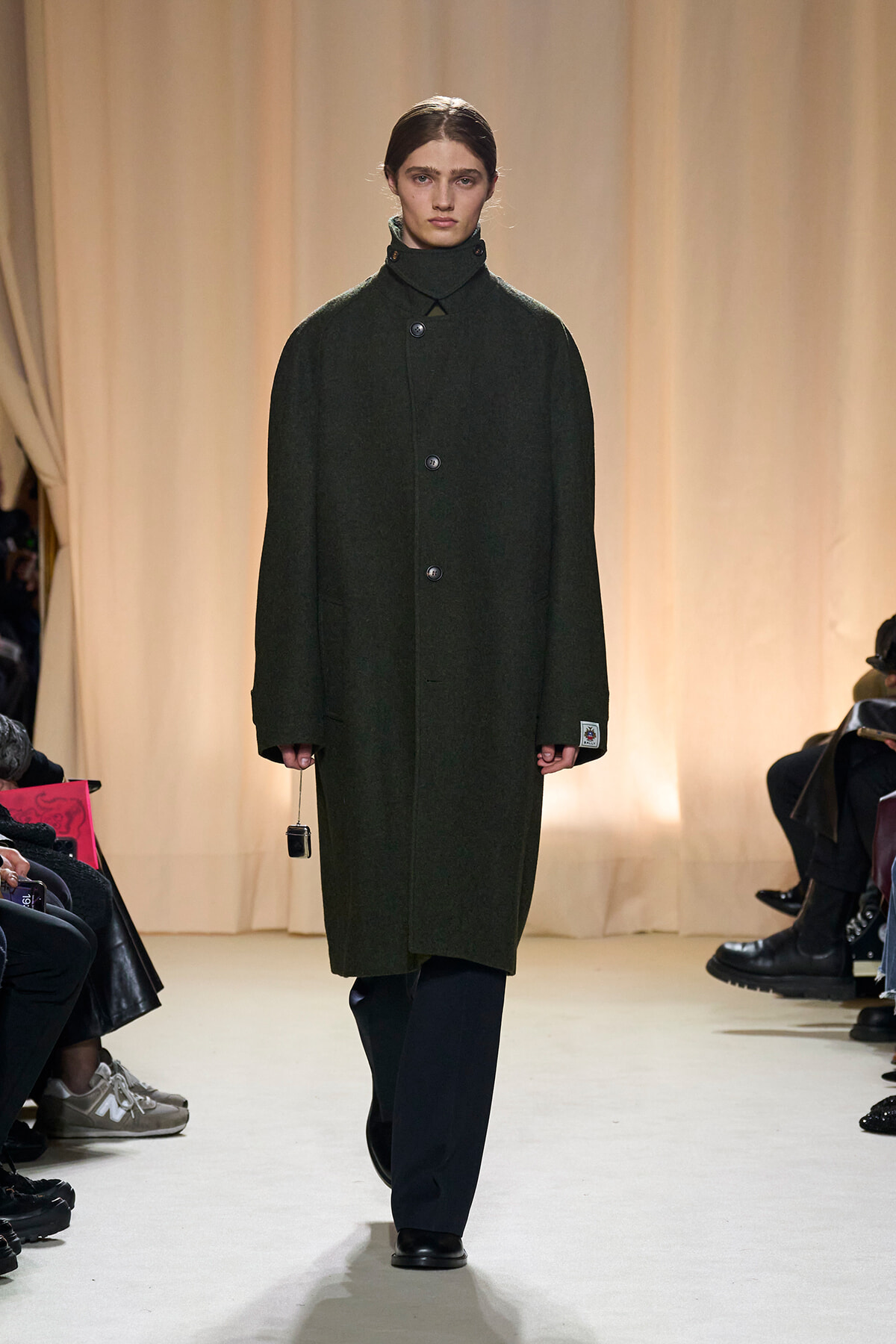 Model walking a runway in a dark green wool overcoat with a high stand collar; audience along the sides.