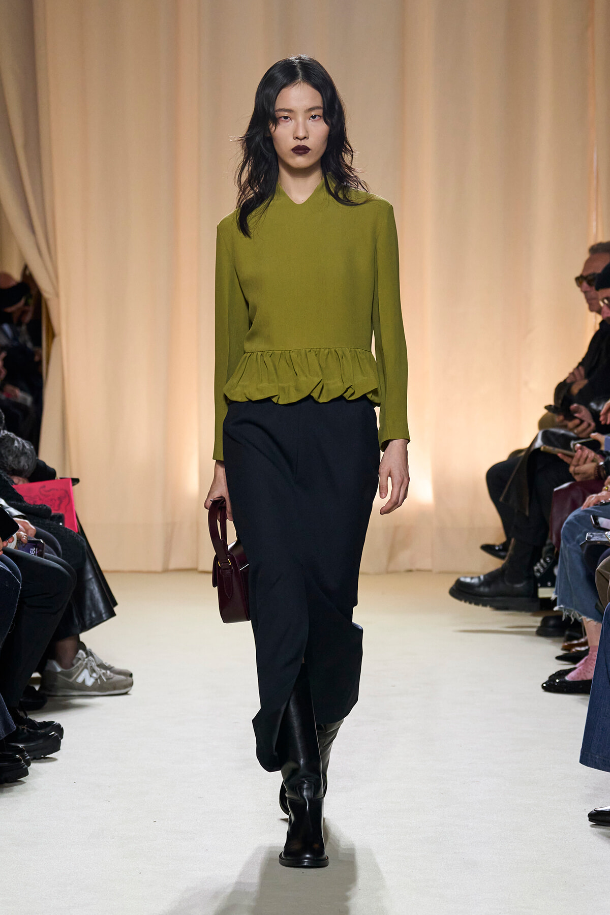 Model walking a runway in an olive-green peplum top and black pants, carrying a burgundy handbag; audience silhouettes on sides awaiting the show.