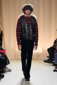 Runway model walking in a red patterned knit sweater under a black leather vest with fur trim on the shoulders.