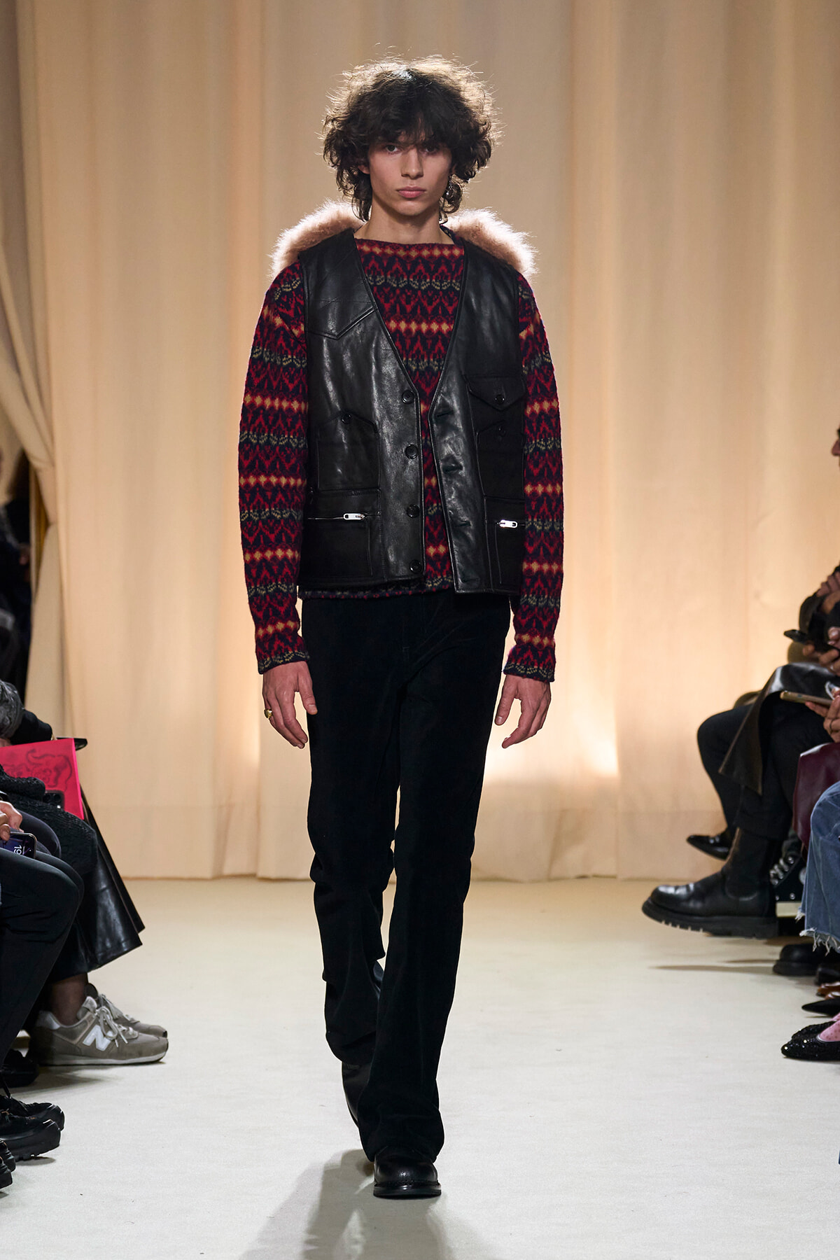 Runway model walking in a red patterned knit sweater under a black leather vest with fur trim on the shoulders.