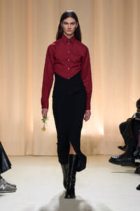 Model walking a runway in a burgundy button-down blouse tucked into a black high-waisted skirt, wearing black boots and holding a small sprig with a gold ornament.