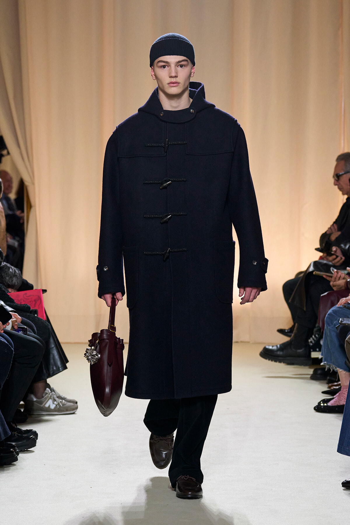 Model walks runway in a long navy duffle coat with toggle closures, beanie, and brown shoes, carrying a burgundy handbag.