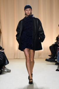 Model on a runway wearing a navy ruffled dress under a black oversized leather coat, with a navy beanie and black shoes.