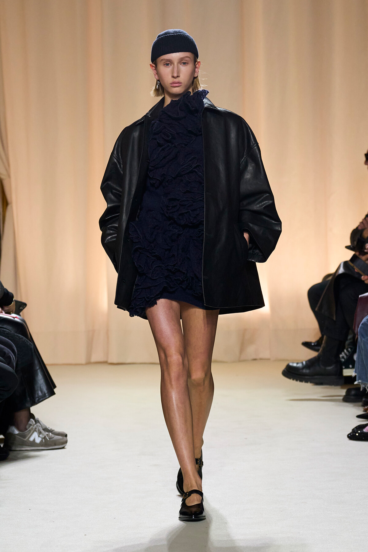 Model on a runway wearing a navy ruffled dress under a black oversized leather coat, with a navy beanie and black shoes.