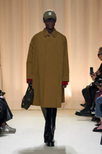 Model walks the runway wearing a camel overcoat, olive cap, dark sunglasses, black trousers, and carries a green handbag; beige backdrop.
