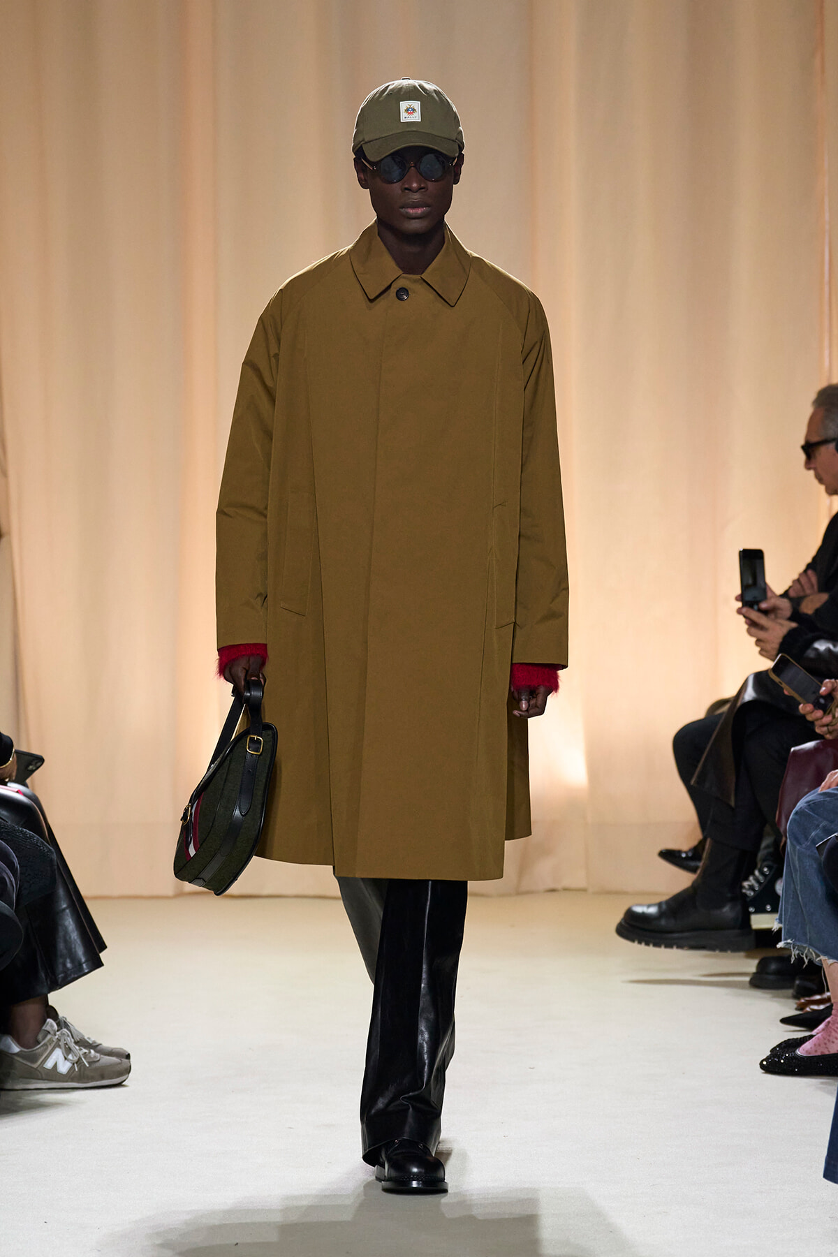 Model walks the runway wearing a camel overcoat, olive cap, dark sunglasses, black trousers, and carries a green handbag; beige backdrop.
