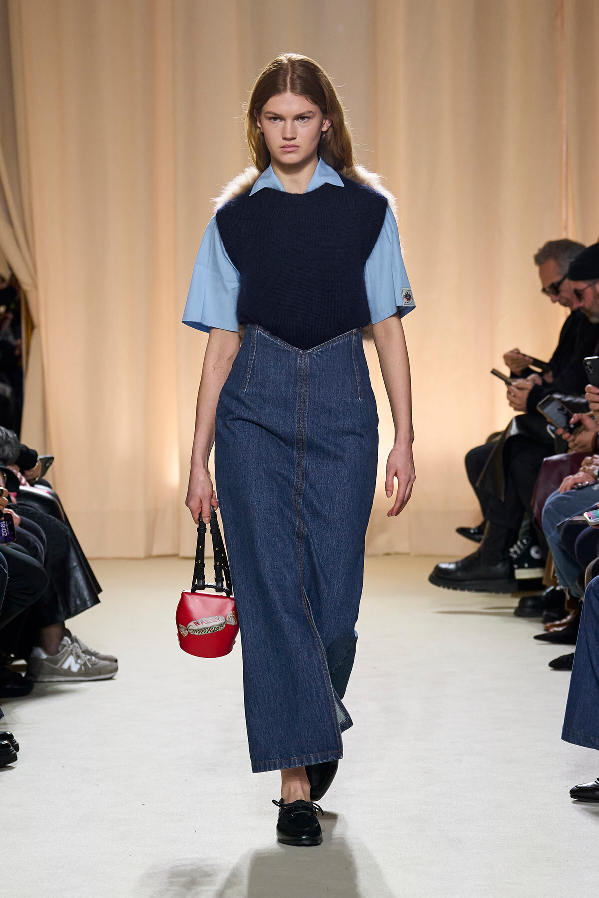 Model walks a runway wearing a light blue shirt under a navy knit vest and a high-waisted denim skirt, carrying a small red handbag.
