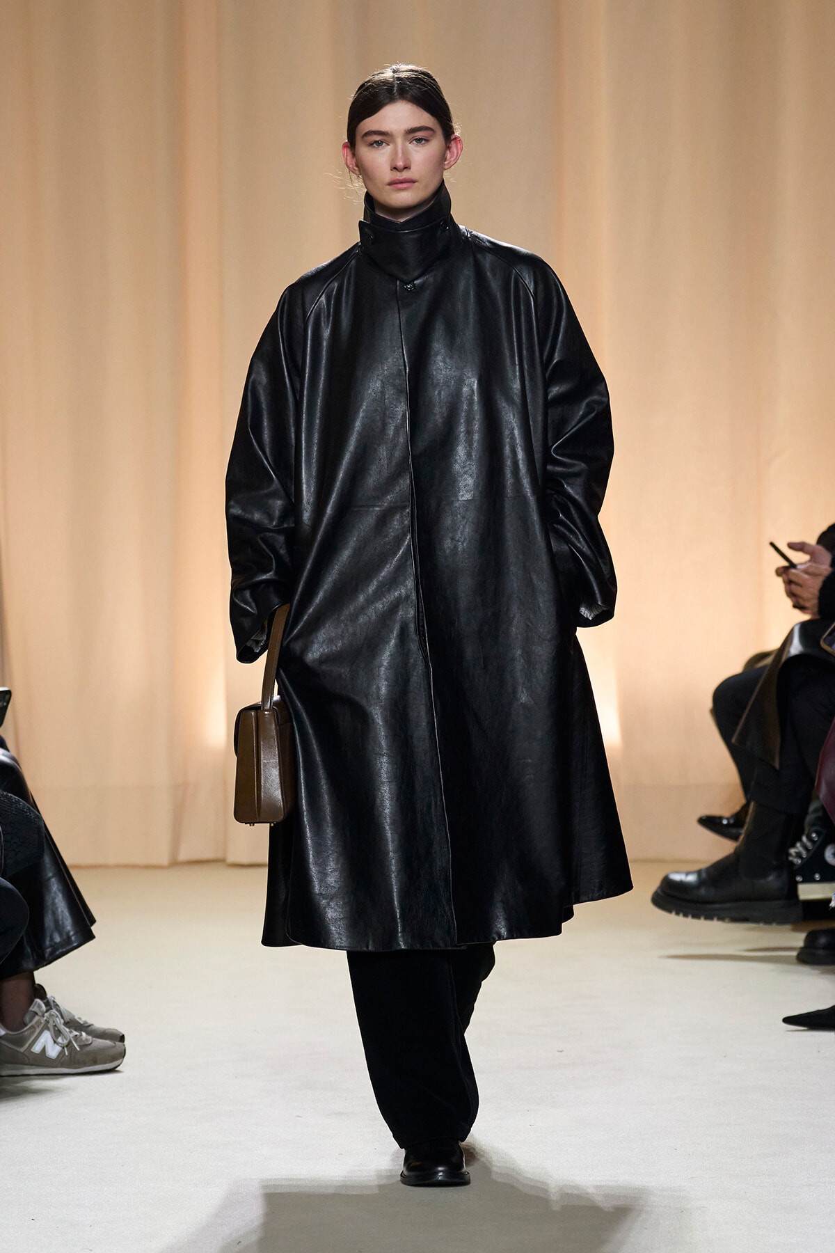 Model walking the runway in a long black leather coat with a high collar, carrying a small brown rectangular bag.