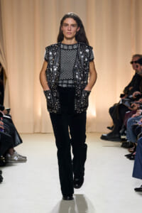 Female model walks a runway wearing a black studded leather vest over a black-and-white geometric sweater, paired with black pants and boots, with an audience on both sides.