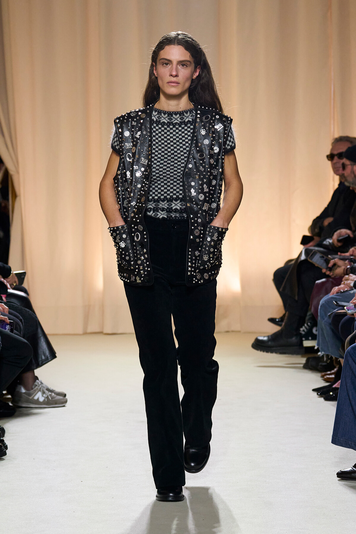 Female model walks a runway wearing a black studded leather vest over a black-and-white geometric sweater, paired with black pants and boots, with an audience on both sides.