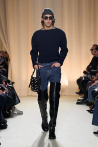 Male model walking the runway in a dark knit sweater, blue jeans, black knee-high boots, gray beanie, round sunglasses, and a studded black handbag.
