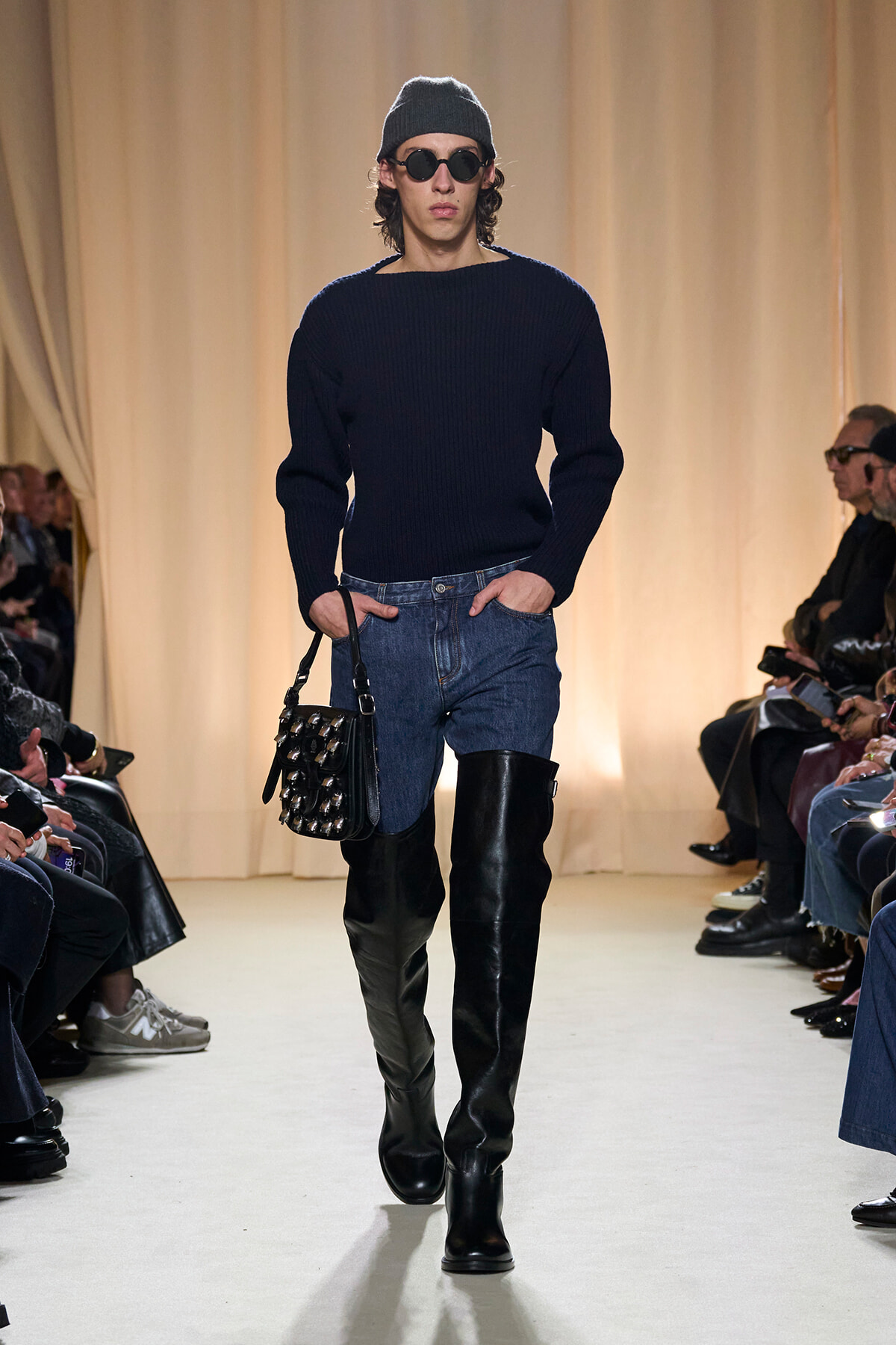 Male model walking the runway in a dark knit sweater, blue jeans, black knee-high boots, gray beanie, round sunglasses, and a studded black handbag.