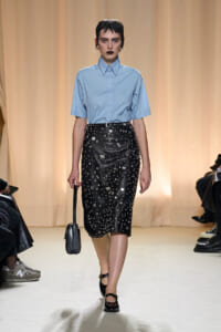 Model on a runway wearing a light blue short-sleeve button-down shirt with a black studded midi skirt and a small black handbag.