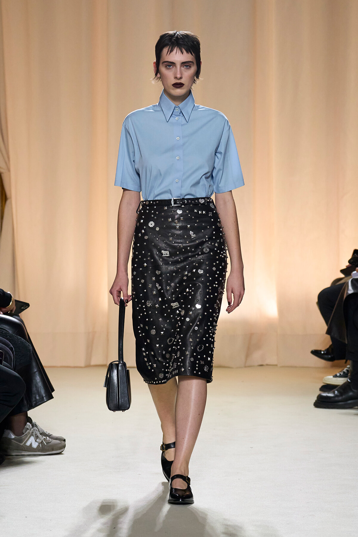 Model on a runway wearing a light blue short-sleeve button-down shirt with a black studded midi skirt and a small black handbag.