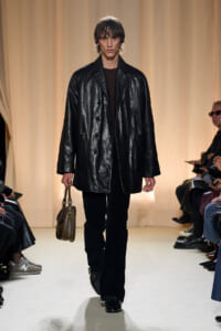 Male model walks a fashion runway in a shiny black leather overcoat, dark sweater, black pants, and a brown satchel at his side.