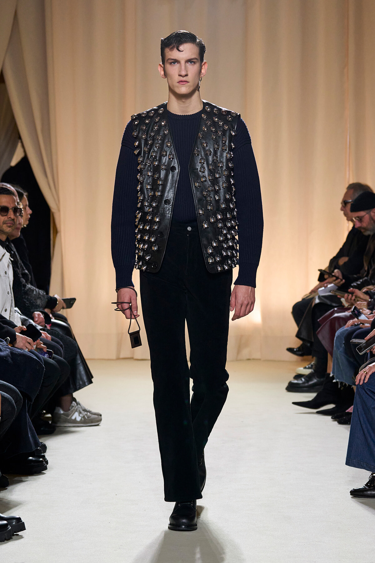 Male model walking the runway in a navy knit sweater and a black studded leather vest, with black trousers and sunglasses in hand.