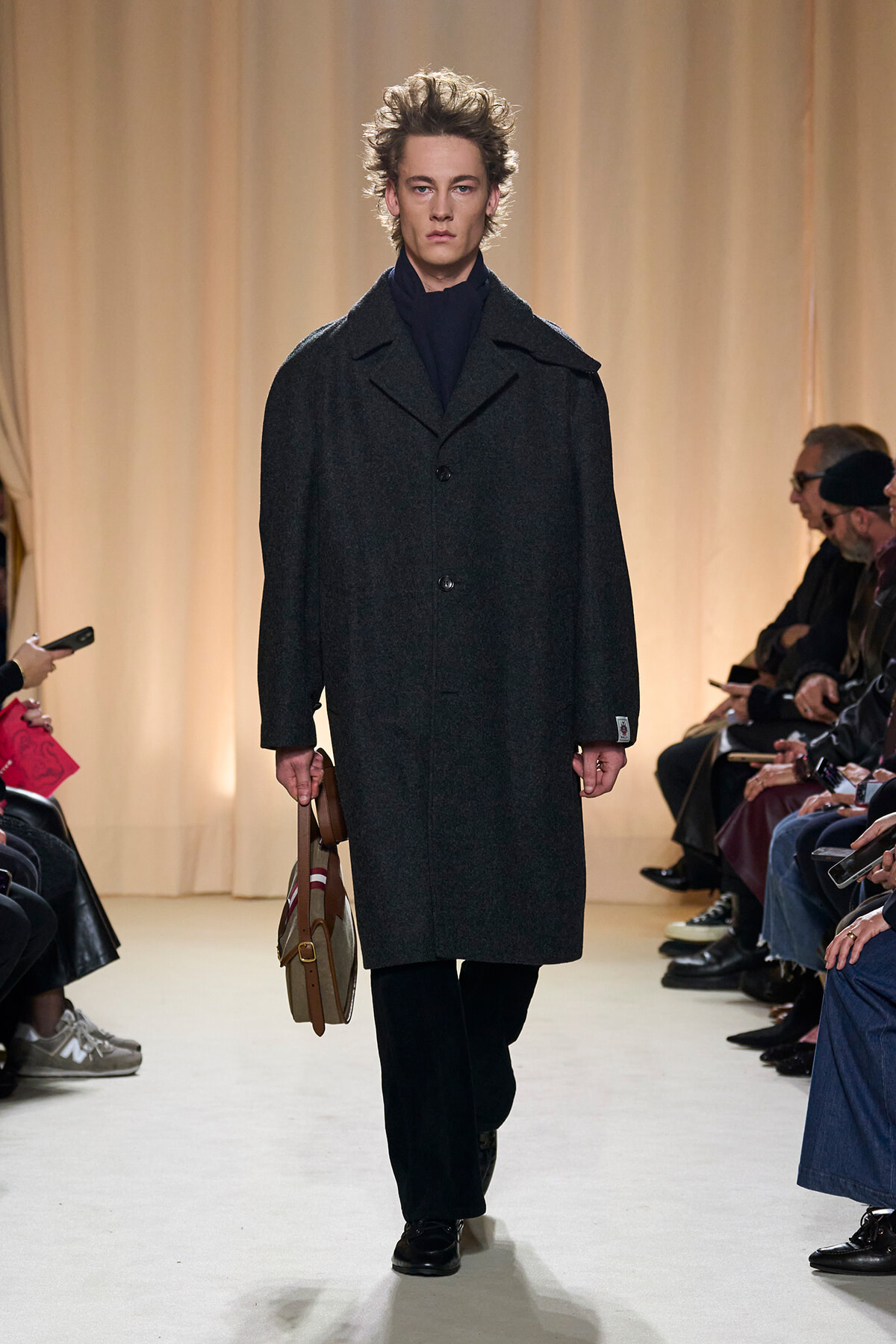 Male fashion model walking a runway in a dark wool overcoat with a navy scarf, carrying a tan leather bag, audience seated along the sides.