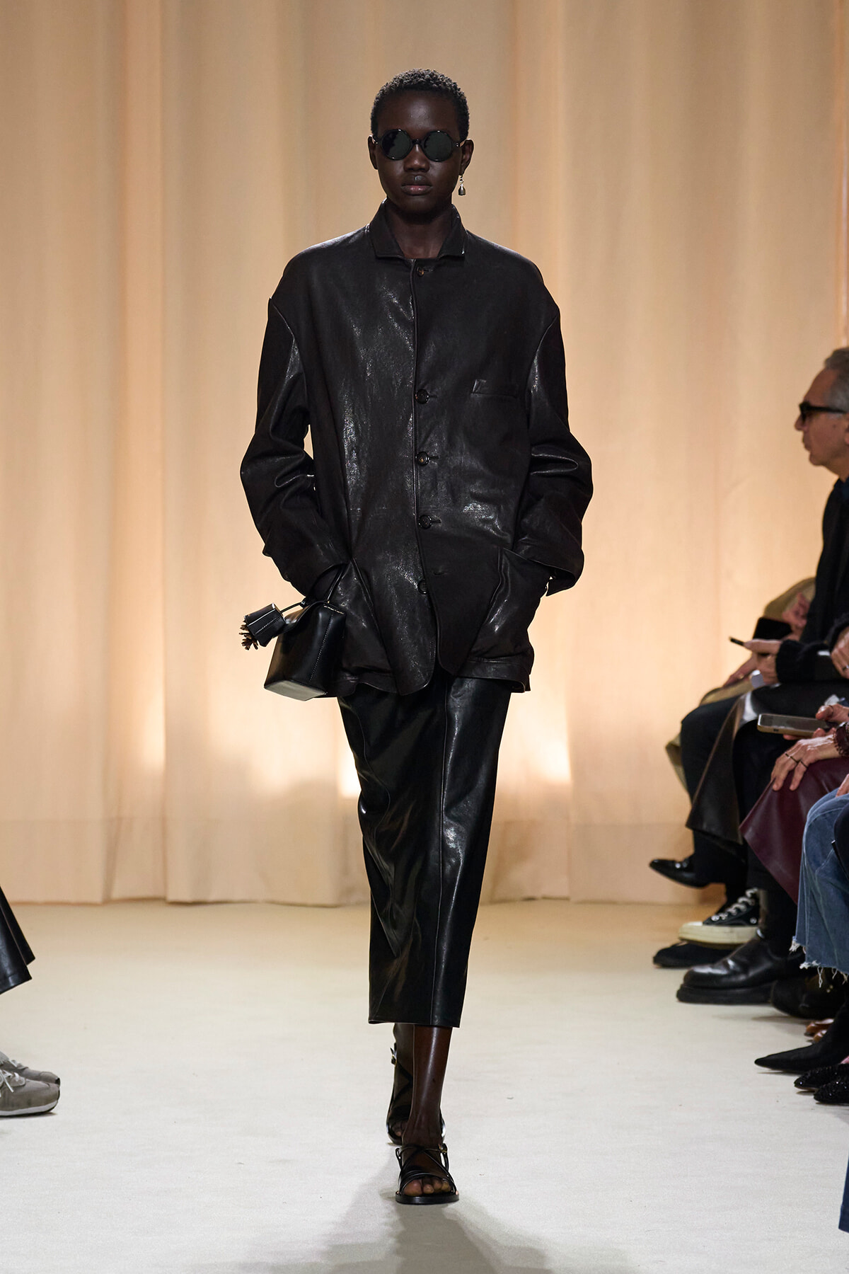 Runway model walking in a shiny black leather coat and matching pants, wearing dark sunglasses and carrying a small black bag.