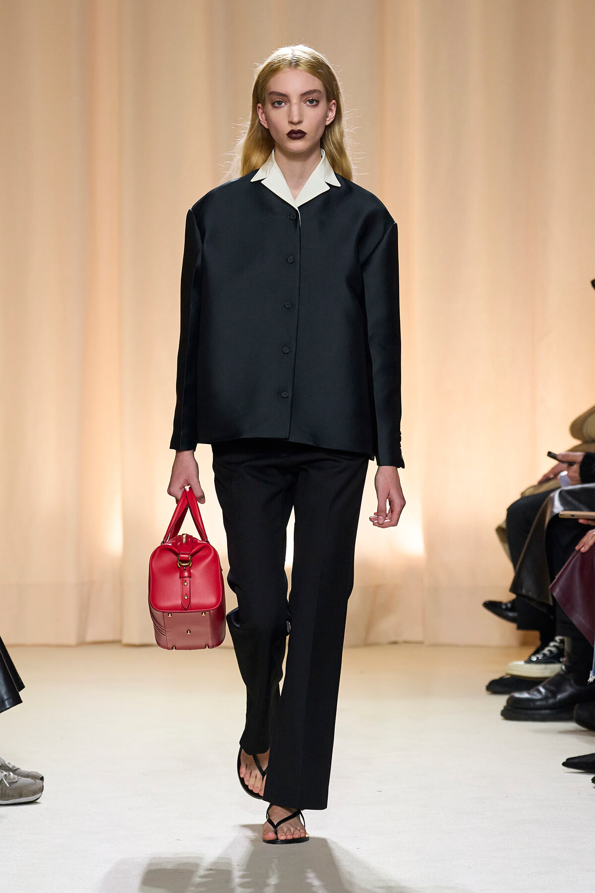Model walking a fashion runway in a black suit with a white collared shirt, holding a bright red handbag and wearing black flip-flop sandals against a warm beige backdrop.
