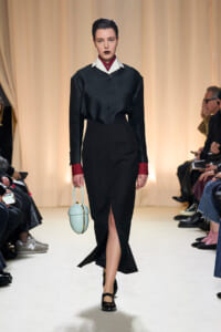 Model walking a runway in a black dress with a white collar and red cuffs, carrying a pale blue rounded handbag; audience seated along the sides in the background.