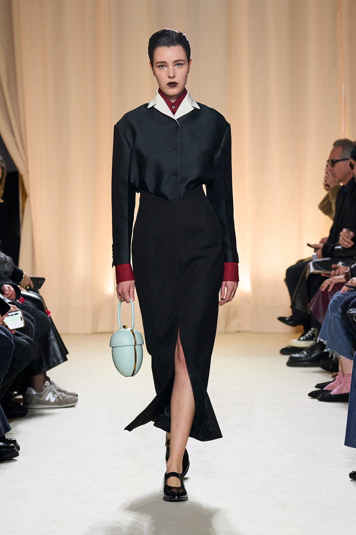 Model walking a runway in a black dress with a white collar and red cuffs, carrying a pale blue rounded handbag; audience seated along the sides in the background.