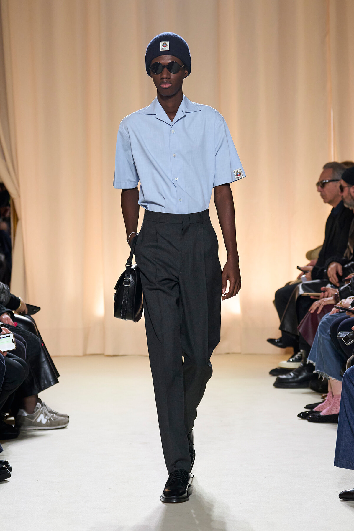 Male model on a runway wearing a light blue short-sleeve shirt, dark pleated trousers, black beanie, sunglasses, and a black satchel.