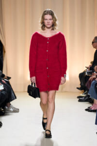 Model walks the runway in a bright red fuzzy cardigan buttoned down the front, carrying a small black handbag.